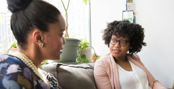 Two Black women sitting on a couch in a white room, in front of a windowsill covered in plants, in deep discussion.