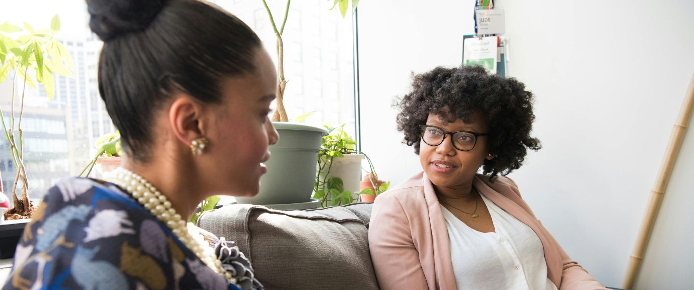 Two Black women sitting on a couch in a white room, in front of a windowsill covered in plants, in deep discussion.
