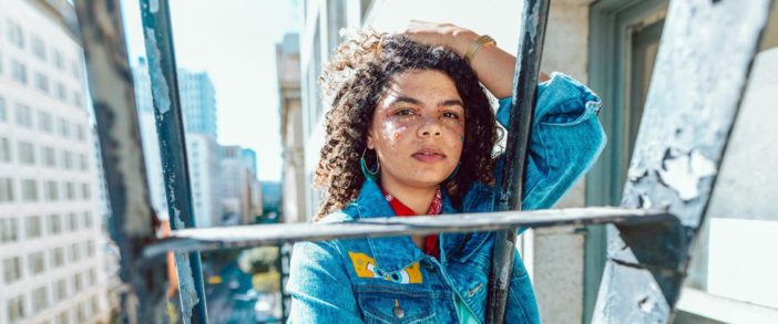 A femme presenting Black person with stick on gems on their face and wearing a denim jacket, holding their curly hair back with their hand, standing on a fire escape in a city.