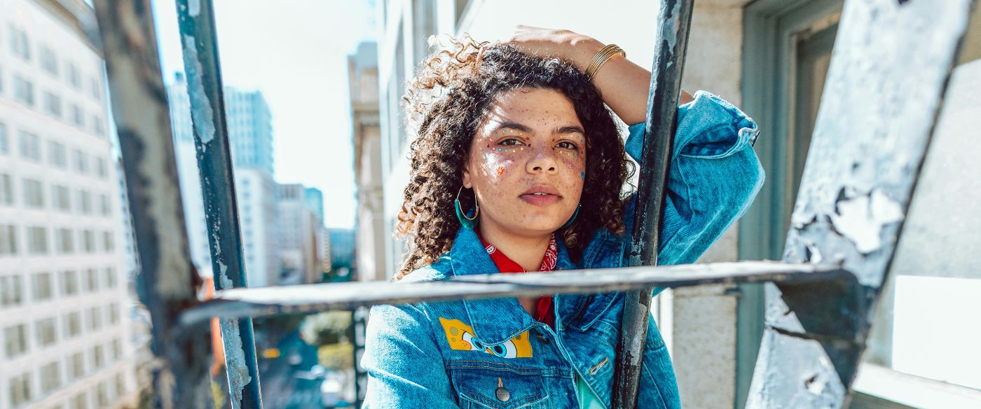 A femme presenting Black person with stick on gems on their face and wearing a denim jacket, holding their curly hair back with their hand, standing on a fire escape in a city.